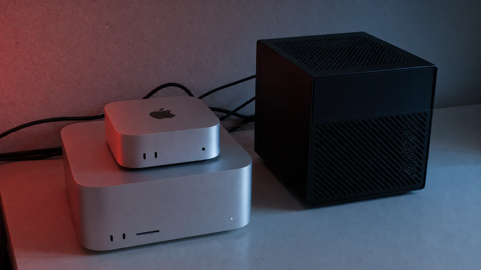 A wall shelf with Mac minis, Mac Studios, a NAS and a network switch in red and blue duotone light