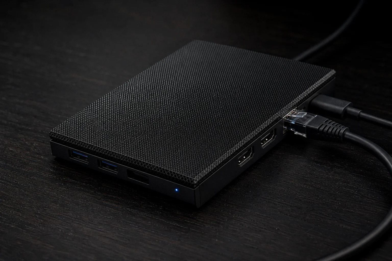 A fanless mini PC with ethernet and USB-C connected, sitting on a dark desk with a single blue status LED